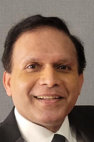 A man with short black hair, wearing a black suit and white shirt, smiling.