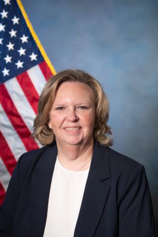A woman in a navy blazer and white shirt smiles in front of an American flag.