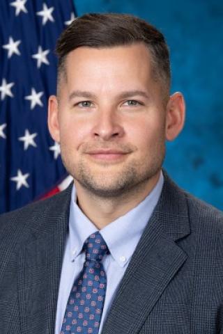 Man in business attire smiles in front of American flag.
