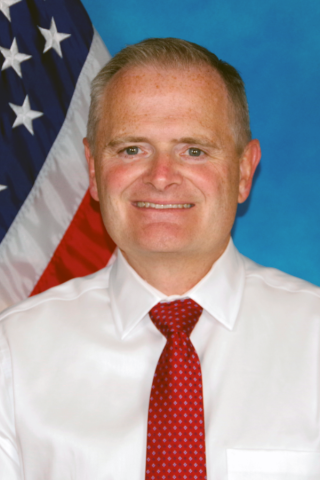 Man in white shirt and red tie in front of an American flag.