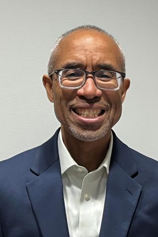 Smiling man in blue blazer over white shirt with glasses and short hair.