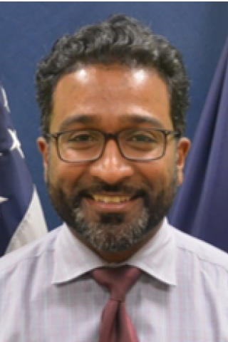 Man with glasses, beard, and red tie smiling in front of US flag.