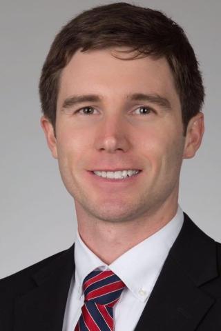 A man in a black suit and red, white, and blue striped tie smiles.