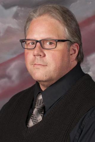 A man in a black shirt, sweater, and tie with an American flag background.