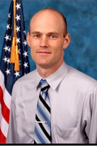 Man with gray shirt, blue tie, and American flag in background.