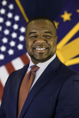 Man in a navy blue suit, red tie, smiling in front of the US flag.