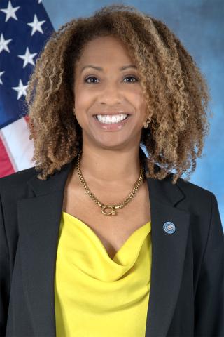 Smiling woman in yellow top and black blazer with US flag in background.
