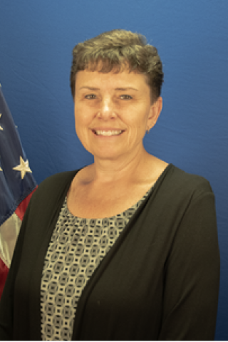 Woman smiling with short hair wearing a black cardigan and patterned shirt next to an American flag.