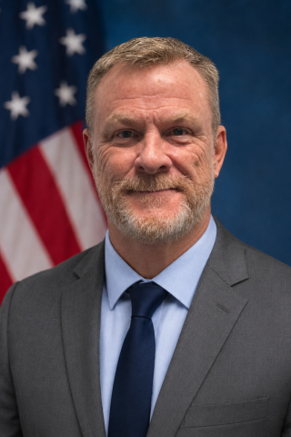 Man in business attire stands in front of American flag, smiling.