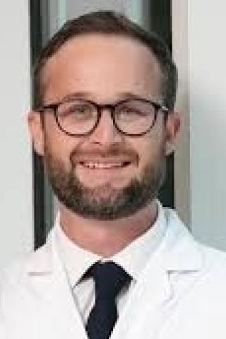 Smiling man in white lab coat and black tie stands near window.
