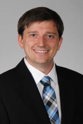 Man in black suit and blue plaid tie smiling for the camera.