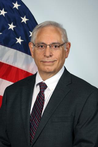 Man in a dark suit, red-striped tie, and glasses, standing in front of an American flag.