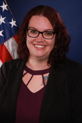 Woman with glasses and maroon top smiles in front of American flag.