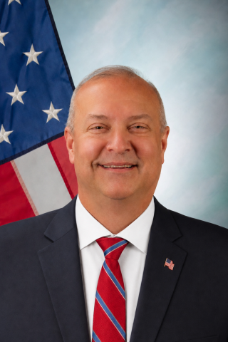 Man in suit and tie smiling with American flag in background.