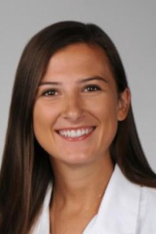 Woman with long brown hair smiling, wearing a white lab coat.