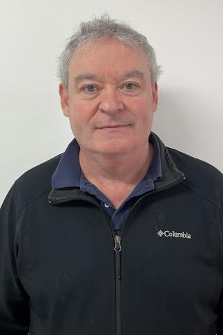 Man with gray hair, wearing black jacket and blue shirt, smiling against white background.