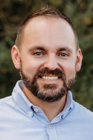 Headshot of man with beard smiling, wearing light blue button-up shirt.