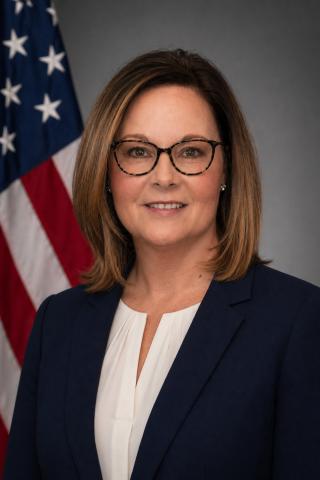 Portrait of a woman in a blue blazer and white blouse, standing before an American flag.