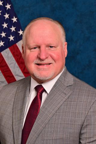 Smiling man in a suit and tie in front of an American flag.