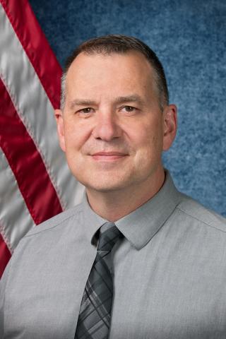 Man in gray shirt and tie standing in front of American flag.