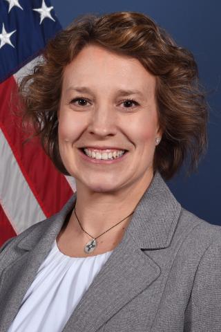 Smiling woman in gray blazer and white shirt with American flag in background.