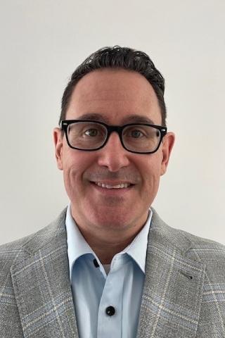 Man in gray plaid blazer and blue shirt, wearing glasses, smiling at the camera.