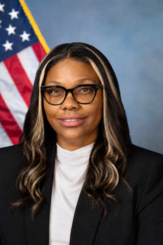 A smiling woman with long hair wearing glasses and a black blazer with an American flag behind her.
