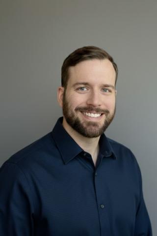 Man with short brown hair and goatee, wearing a navy button-up shirt, smiling against a gray background.