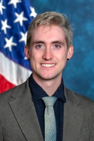 Caucasian man in gray suit, black shirt, and light blue tie smiling in front of an American flag.