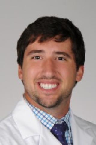 A man in a white lab coat and blue tie smiles.