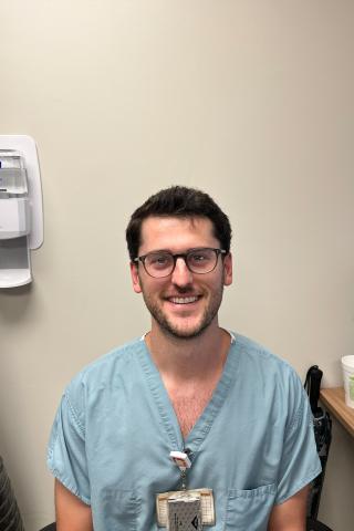 Smiling man in blue scrubs stands in a medical office with a hand sanitizer dispenser on the wall.
