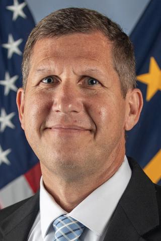 Man in a suit and tie, smiling in front of U.S. and DHS flags.