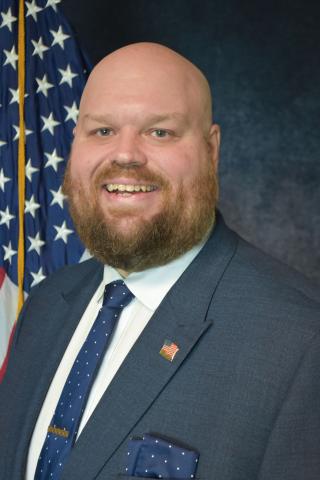 A bald man with a beard in a blue suit smiles in front of an American flag.