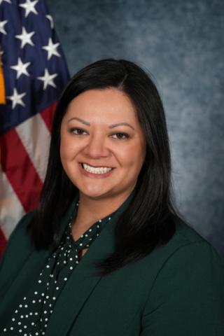 Woman in green blazer and polka-dot scarf smiles in front of American flag.