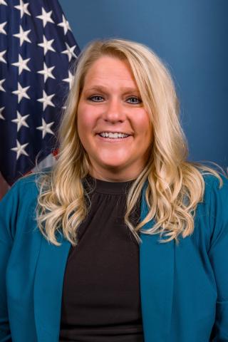 Blonde woman smiling, wearing a blue jacket and black top, with an American flag in the background.