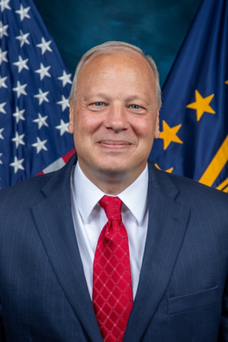 Man in a blue suit and red tie stands in front of American and state flags.
