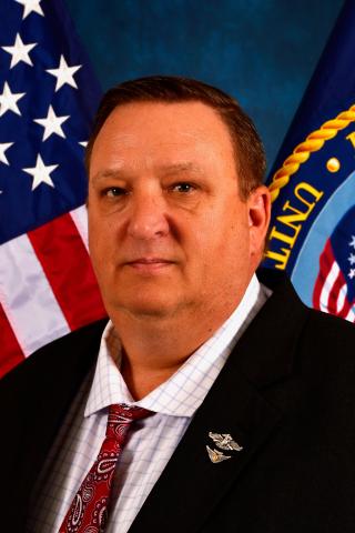 Man in suit and tie standing before American and U.S. Department of Defense flags.