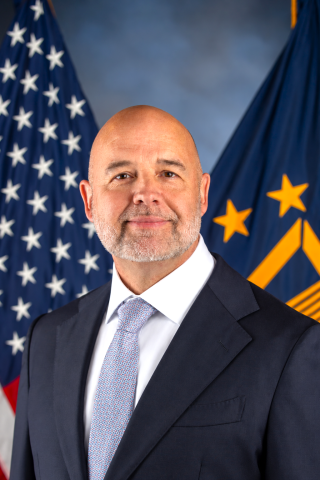 A man in a dark suit and light blue tie stands in front of the American flag.