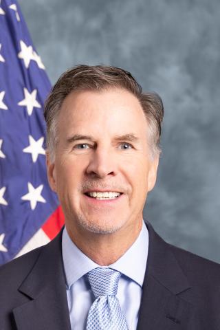 Man in suit and tie smiling, with American flag in background.