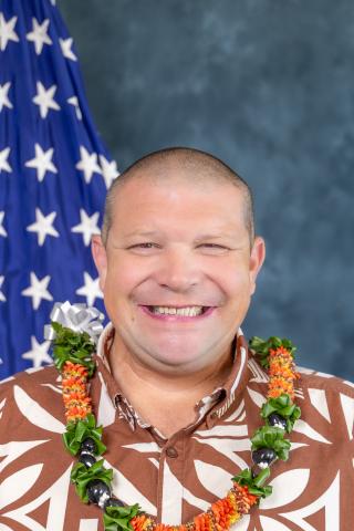 Portrait of a smiling man wearing a lei and standing in front of a flag.