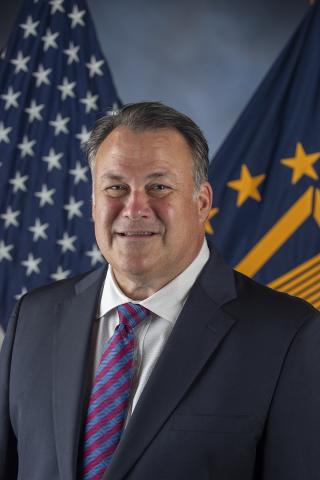 Man in suit and tie standing before an American flag and another flag.
