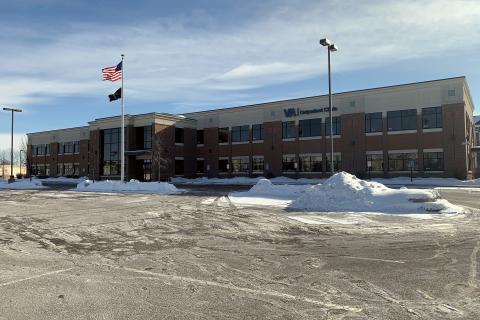 photo of large red brick and tan stucco building with many windows; large snow-filled parking lot in foreground; American and POW flags in front of building