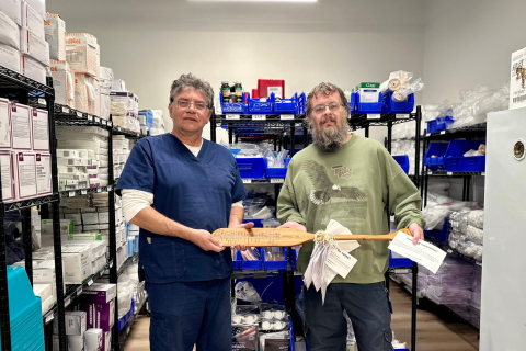 Two men, one in a blue scrub and the other in a green shirt, standing in a storage room holding a paddle.