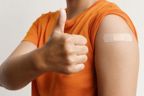 person in orange shirt with bandaid on shoulder and holding a thumbs up.
