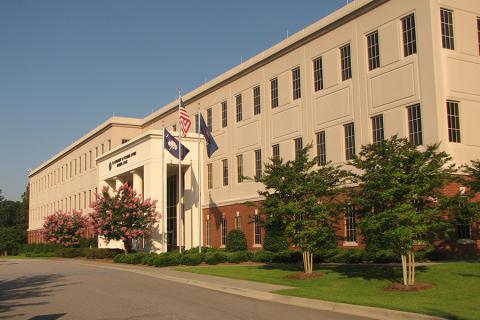 Three story building with large white awning and three flag poles to the right. Bottom of building is brick, top is beige stucco 