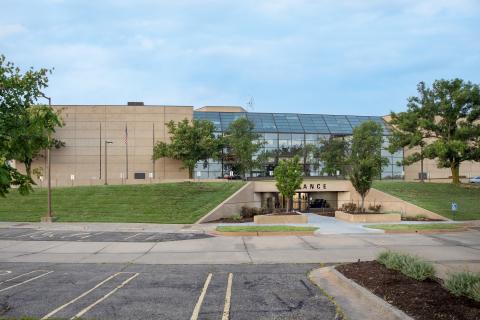 Modern building with glass facade, surrounded by trees and grass, and a parking lot.