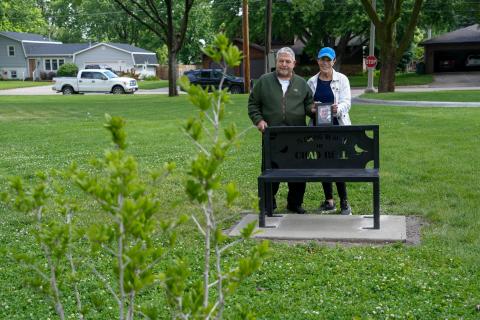 A man and a woman stand behind a black metal bench holding a framed photo. the bench is inscribed with "In Loving Memory of Chad Bell" 
