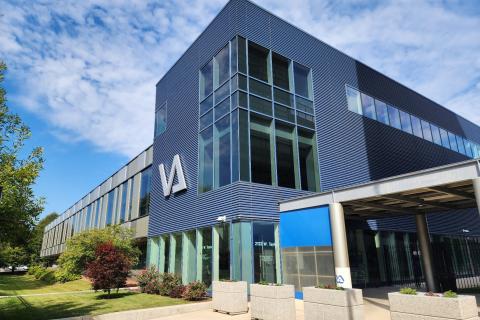 Building enclosed by metal fencing with opening in front. Four spaced concrete plant holders located before the entrance to the right of building.  