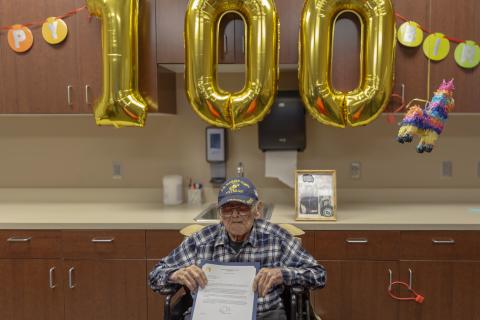 Veteran in wheelchair holds certificate in front of 100th birthday balloons.
