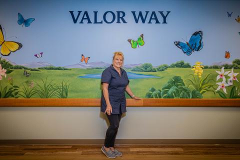 Woman stands in front of mural with butterflies, flowers, and landscape.